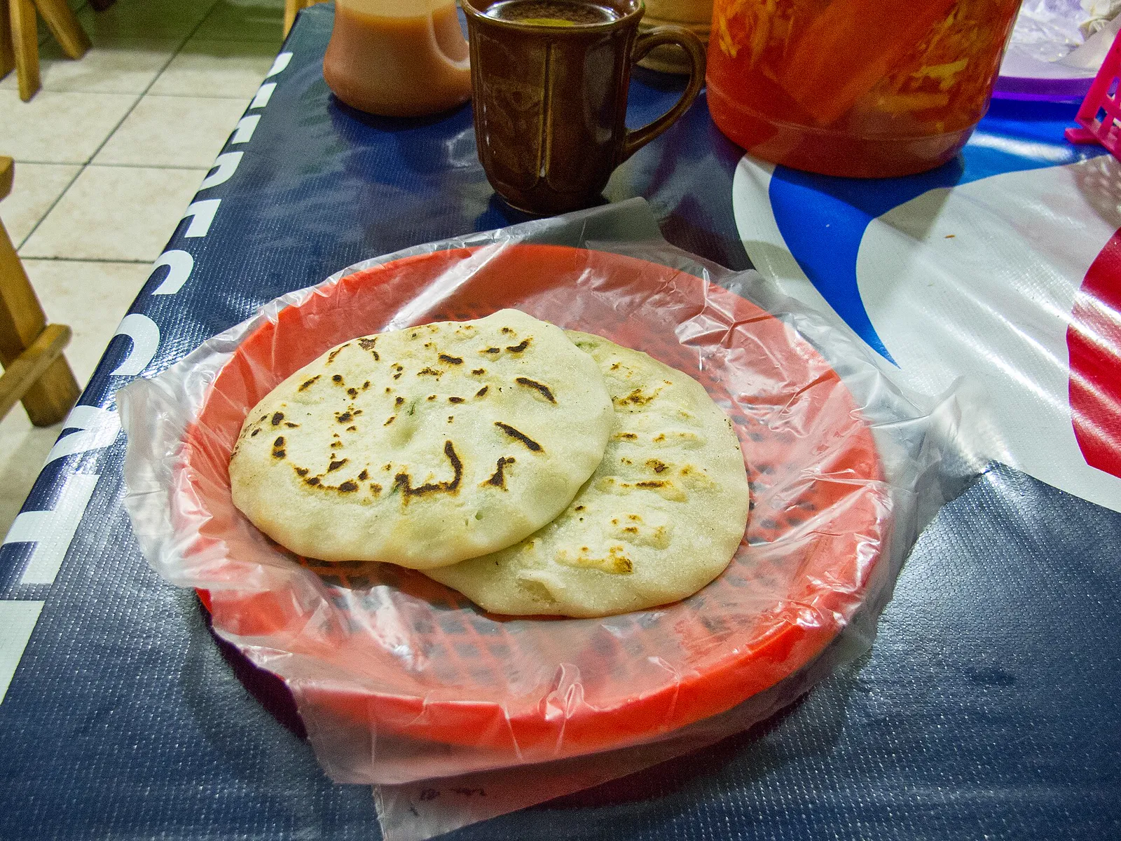 Two pupusas on a red plate, curtido jar behind